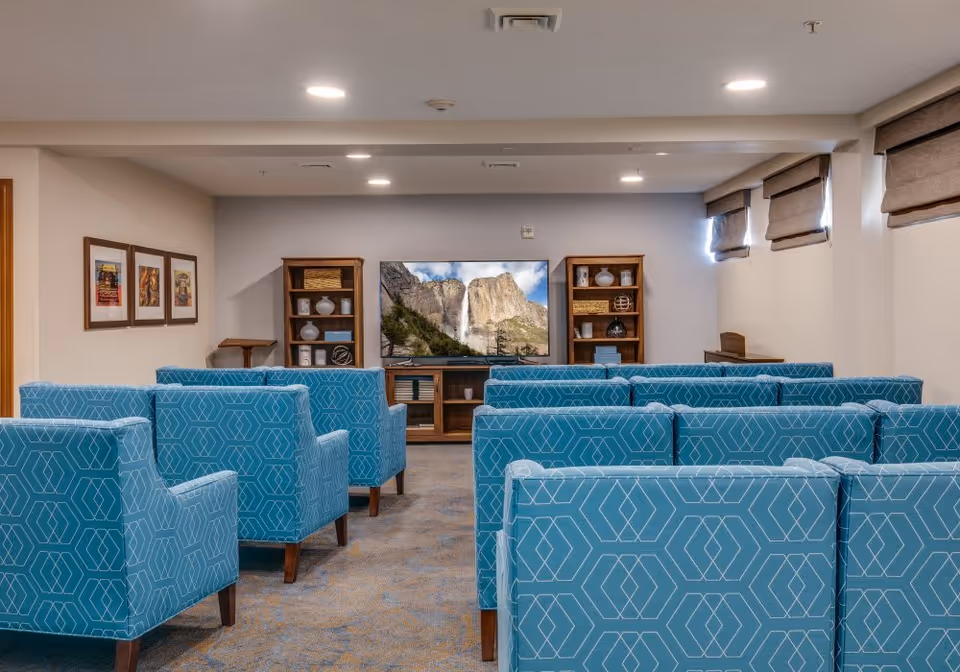 A small theater-style room with rows of blue patterned armchairs facing a large flat-screen TV mounted on a wooden stand. The TV displays an image of a waterfall and rocky cliffs. On either side of the TV are wooden shelves with decorative items. The room has beige walls, carpeted floor, and small windows with brown blinds.