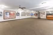 A spacious room with carpeted floor and white walls featuring framed pictures and an American flag display. The room has ceiling fans with lights, a wall-mounted television, and a piano in the corner. The space appears to be a common area or lounge in a senior living facility.