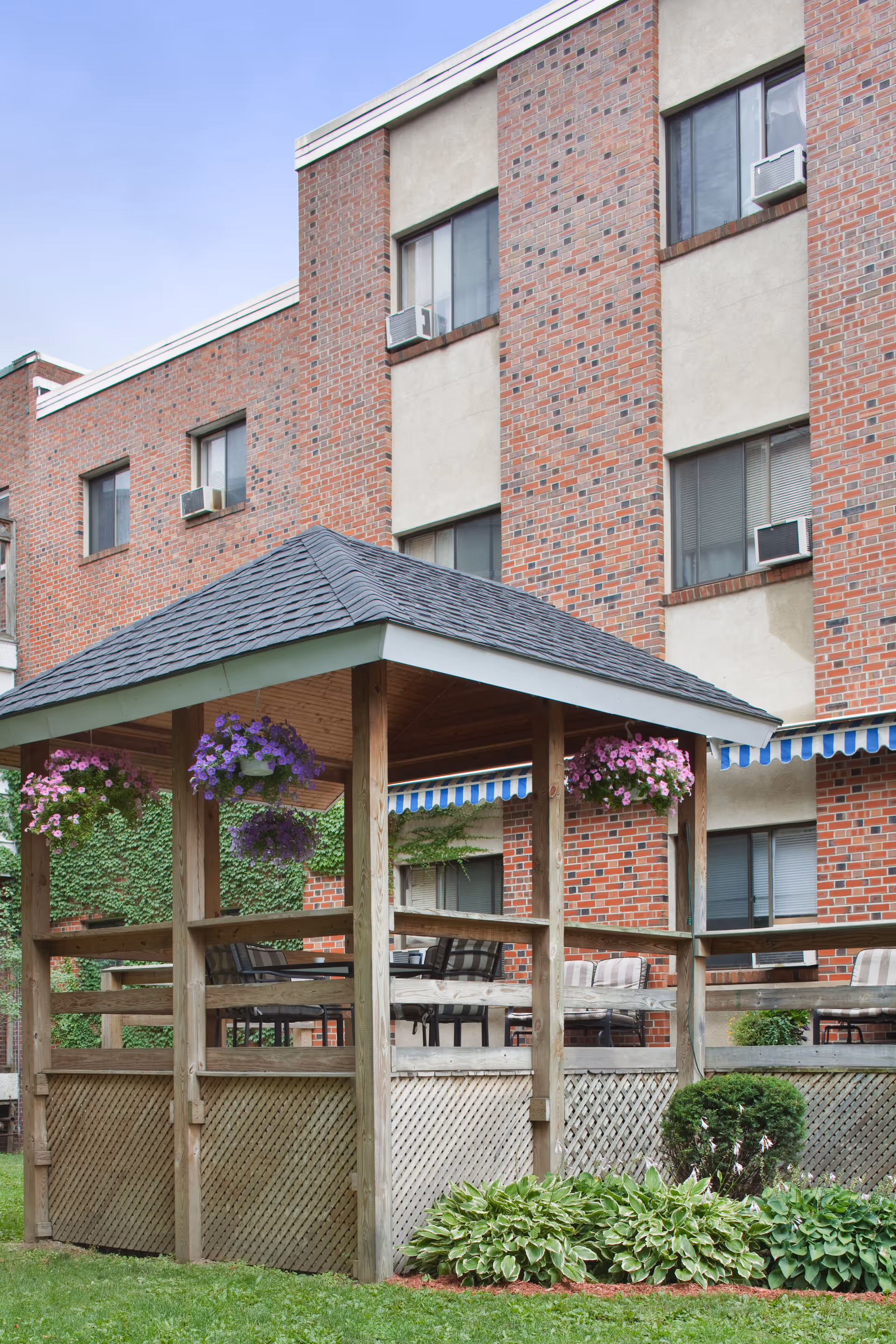 Outdoor wooden gazebo with a shingled roof and hanging flower baskets in front of a multi-story brick building with several windows and window air conditioning units. The gazebo is surrounded by green grass and plants, with outdoor seating visible inside.