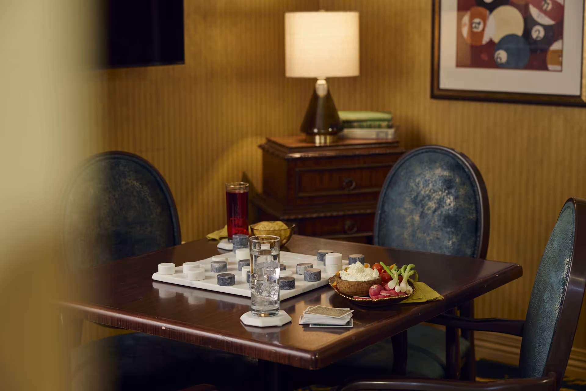 A wooden table with a checkers game set up, two glasses of water, a glass of red beverage, a plate of snacks including radishes and cottage cheese, and a stack of playing cards. The table is surrounded by blue upholstered chairs. In the background, there is a wooden side table with a lamp and some books, and a framed picture on a yellowish wall.