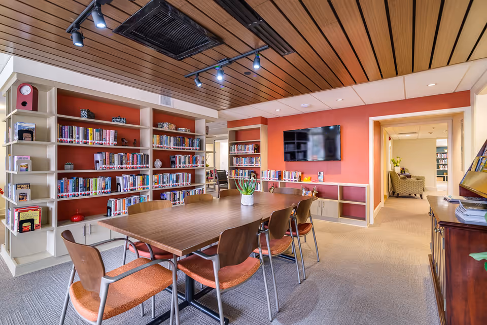 A well-lit library or reading room with a large wooden table surrounded by eight chairs with orange cushions. The room features built-in bookshelves filled with books against a red accent wall, a flat-screen TV mounted on the wall, and a wooden ceiling with track lighting. In the background, there is a hallway leading to another room with a comfortable armchair and additional bookshelves.