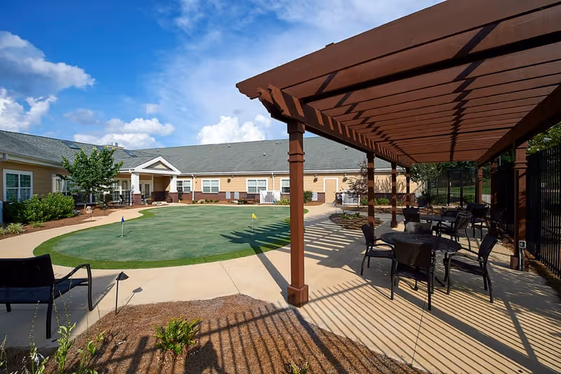 Outdoor patio area at Redstone Village with a wooden pergola casting shadows over several tables and chairs. In the background, there is a putting green surrounded by a paved walkway and single-story building with beige siding and white trim under a blue sky with some clouds.
