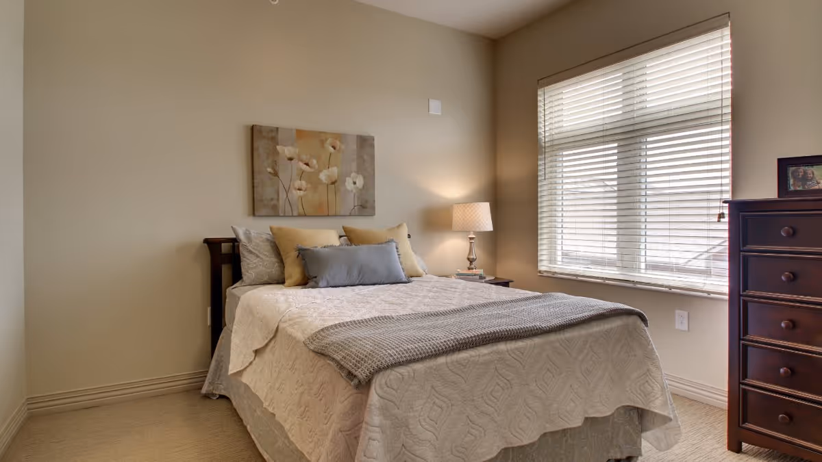 A cozy bedroom with a neatly made bed featuring beige and gray bedding, a wooden headboard, and decorative pillows. A nightstand with a lamp and books is beside the bed, and a window with blinds lets in natural light. A wooden dresser with a framed photo is against the wall.