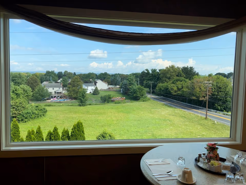 Dining room table set by a large window overlooking a grassy field, road, and nearby houses.