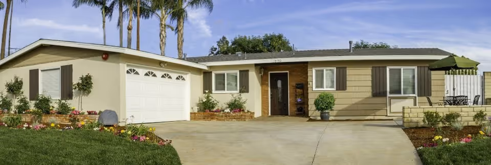 Single-story ranch-style building with an attached two-car garage, driveway, landscaped front yard and palm trees in the background.