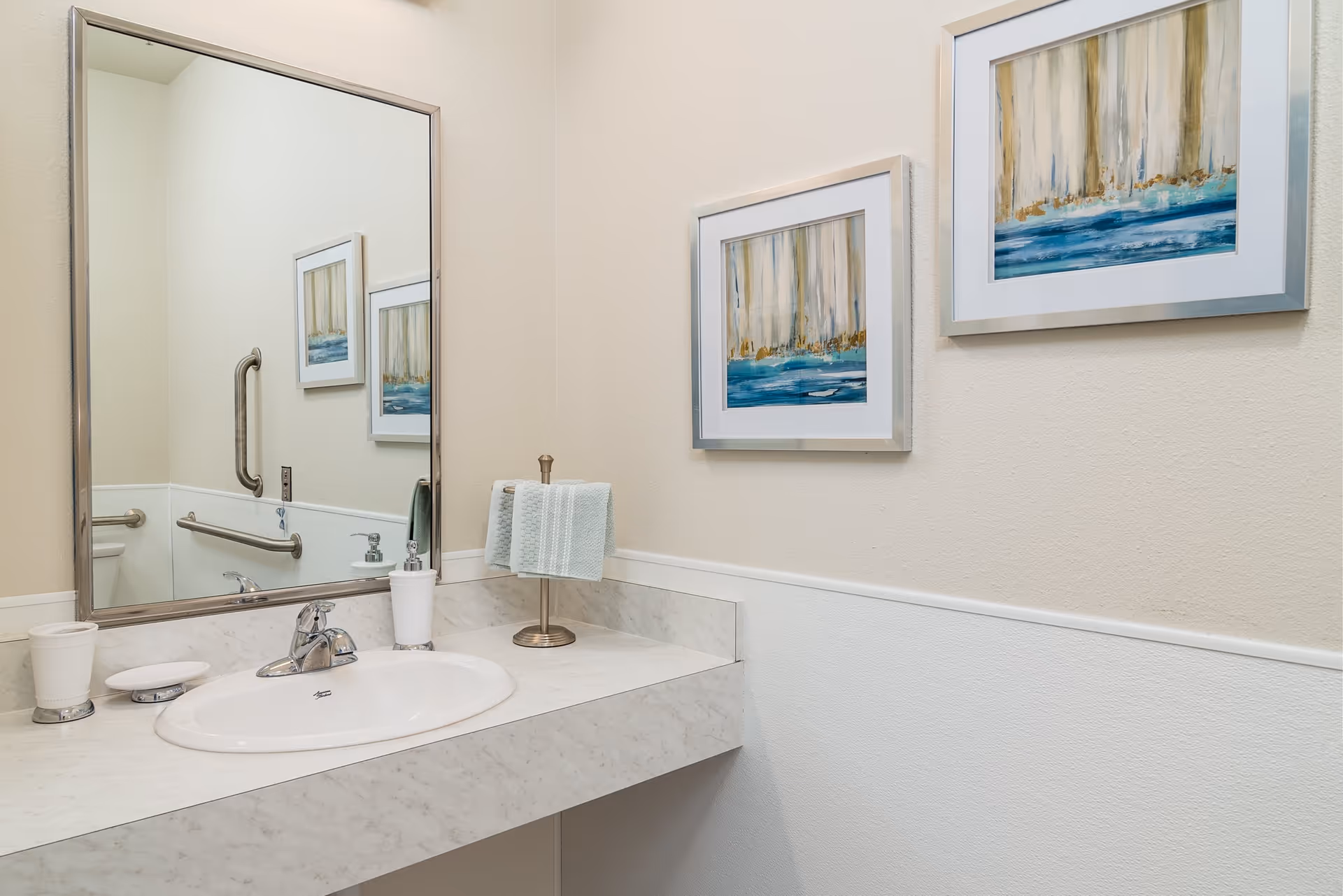 A clean bathroom sink area with a large mirror above it. The countertop is marble with a white oval sink and a chrome faucet. On the counter are a soap dispenser, a small cup, a soap dish with soap, and a towel holder with a folded hand towel. The wall has two framed abstract paintings with blue and beige tones. There are also metal grab bars visible in the mirror reflection.