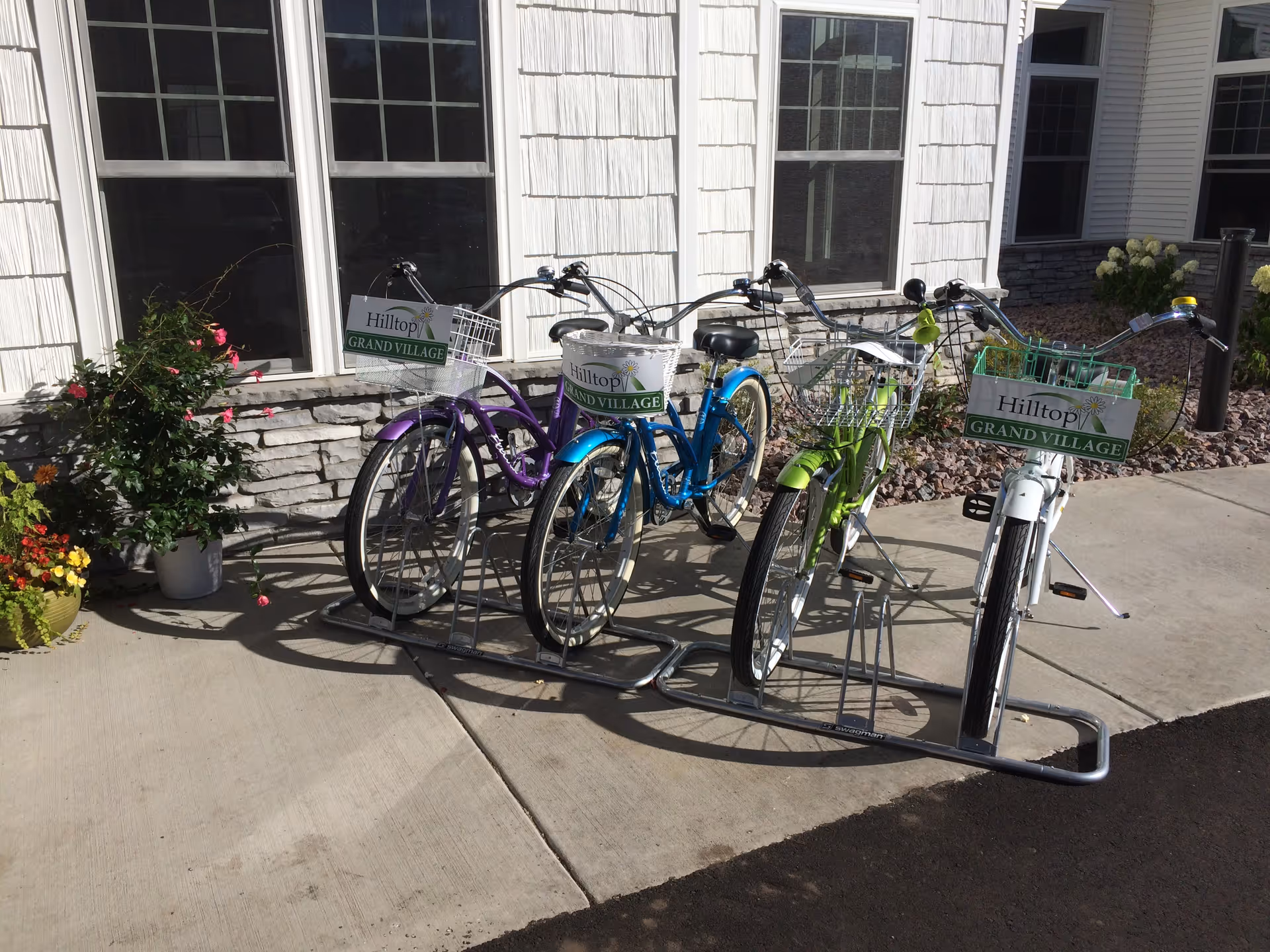 Four colorful bicycles parked in a bike rack outside a building with white siding and stone accents. Each bicycle has a basket with a sign that reads 'Hilltop Grand Village'. There are potted plants with flowers next to the building.