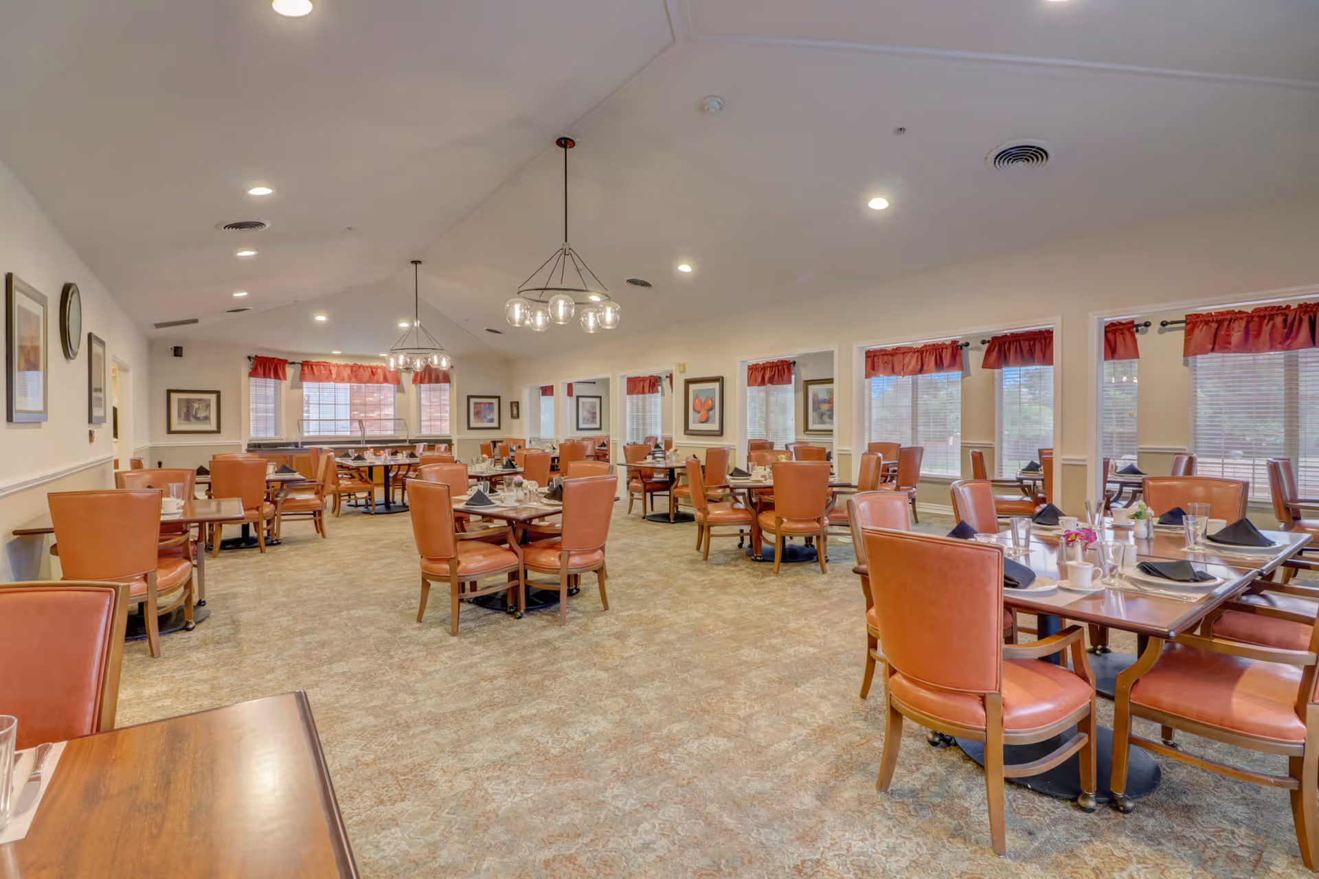 A spacious dining room with multiple wooden tables and orange cushioned chairs arranged neatly. Each table is set with black napkins, glasses, and small flower vases. The room has large windows with red valance curtains allowing natural light to enter. The ceiling is vaulted with modern light fixtures hanging down, and the walls are decorated with framed artwork.