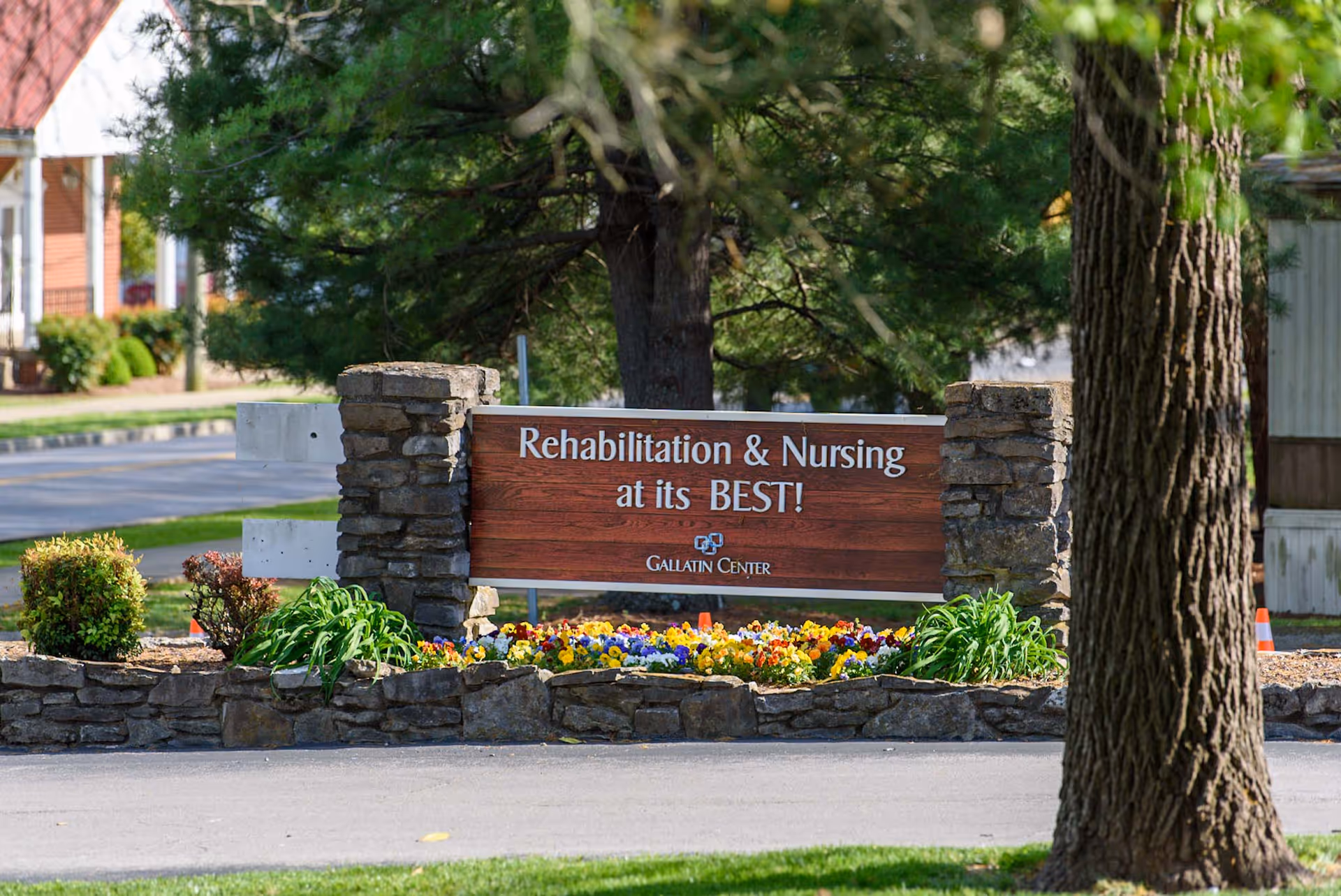 Outdoor view of a wooden sign with stone pillars on each side that reads 'Rehabilitation & Nursing at its BEST! Gallatin Center' surrounded by colorful flowers and greenery with trees and a building in the background.