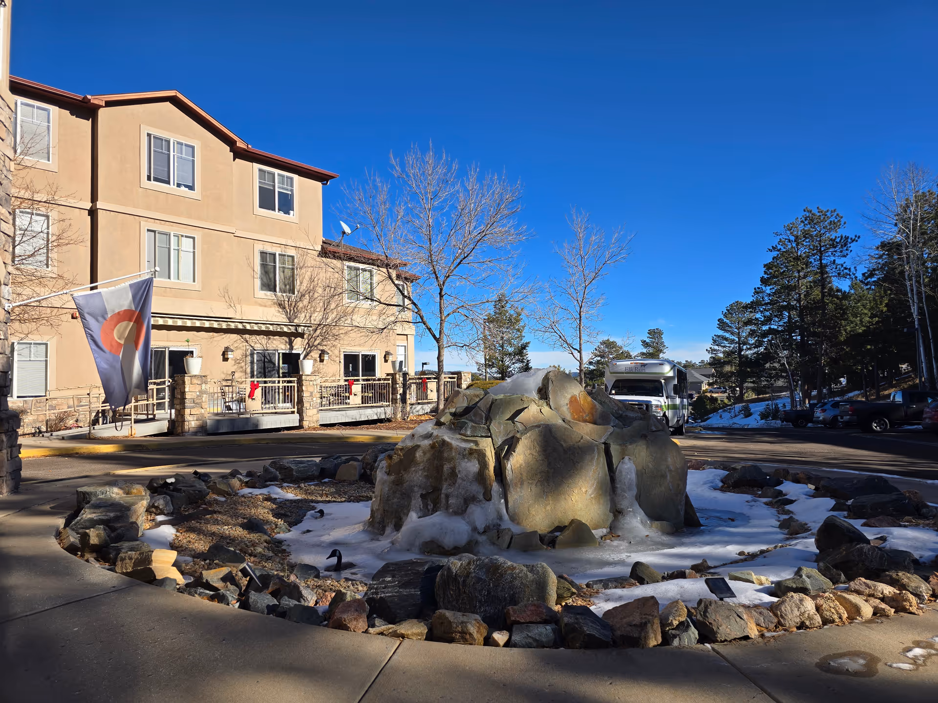 Front exterior of Elk Run Assisted Living with a frozen rock fountain, a flag, parked vehicles, and a clear blue sky.