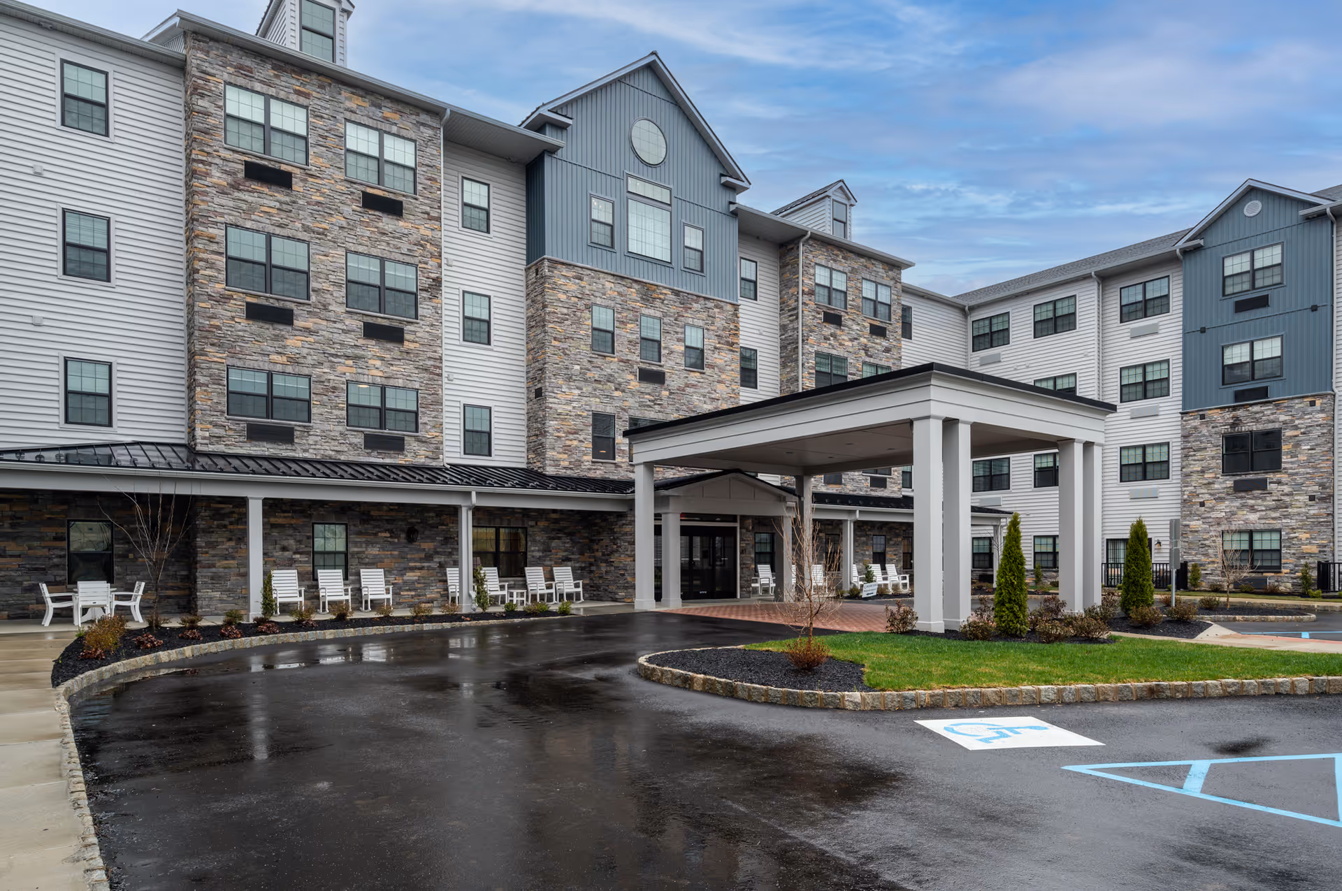 Front exterior view of a multi-story senior living facility with stone and siding facade, a covered entrance with white pillars, several white rocking chairs along the front porch, and a wet driveway with a handicapped parking space visible.