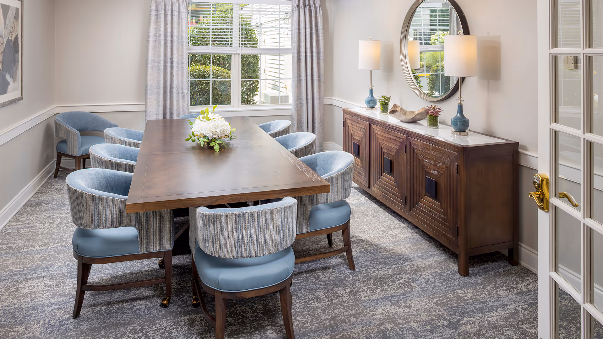 A bright dining room with a long wooden table surrounded by eight blue upholstered chairs. A floral centerpiece is placed on the table. The room features a large window with light curtains, a wooden sideboard with decorative items and two lamps, and a round mirror above the sideboard. The floor is carpeted with a patterned design.