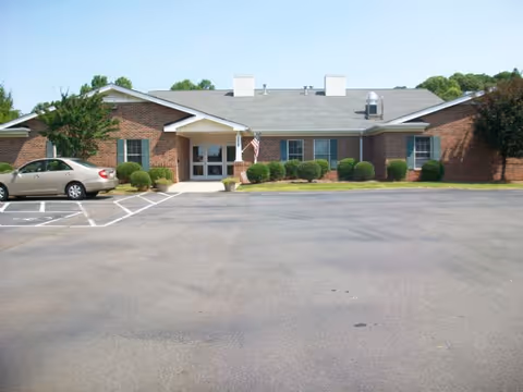 Front exterior view of a single-story brick building with a gray roof, an American flag near the entrance, a parked car in a handicapped parking space, and some bushes and trees around the building.