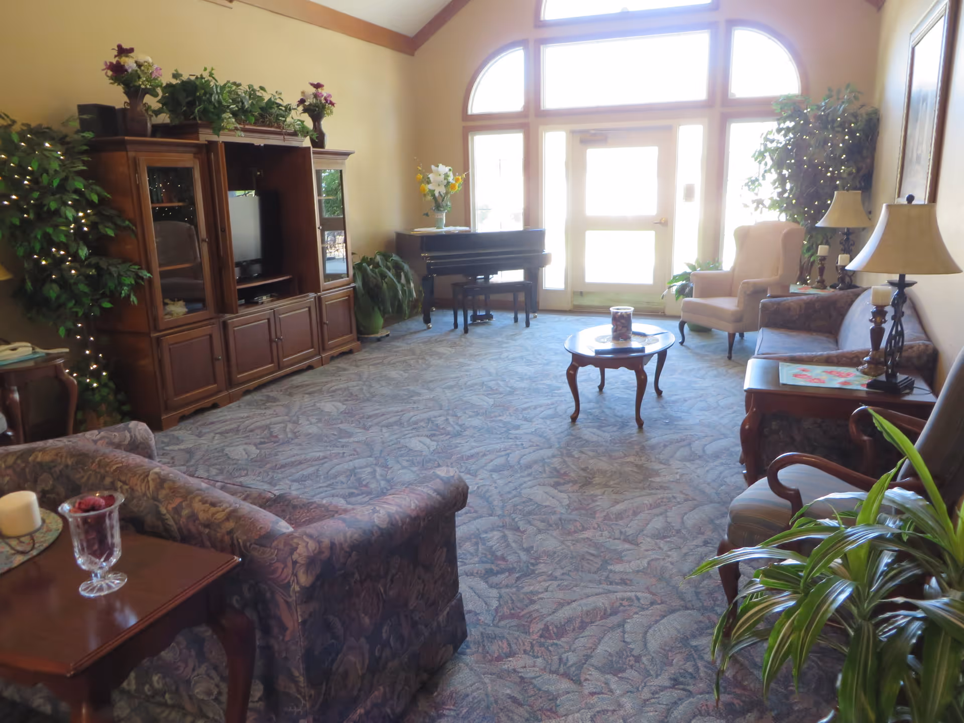 A cozy living room with floral patterned carpet and furniture including sofas, armchairs, and wooden tables. There is a wooden entertainment center with a TV, a black piano near large windows and a door letting in natural light. Several plants and lamps decorate the room.