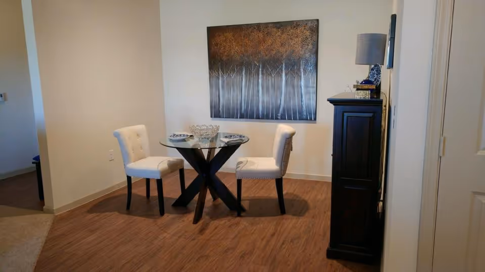 A small dining area with a round glass table and two white upholstered chairs on a wooden floor. A large painting of trees hangs on the wall behind the table. To the right, there is a tall dark wooden cabinet with a lamp and decorative items on top.