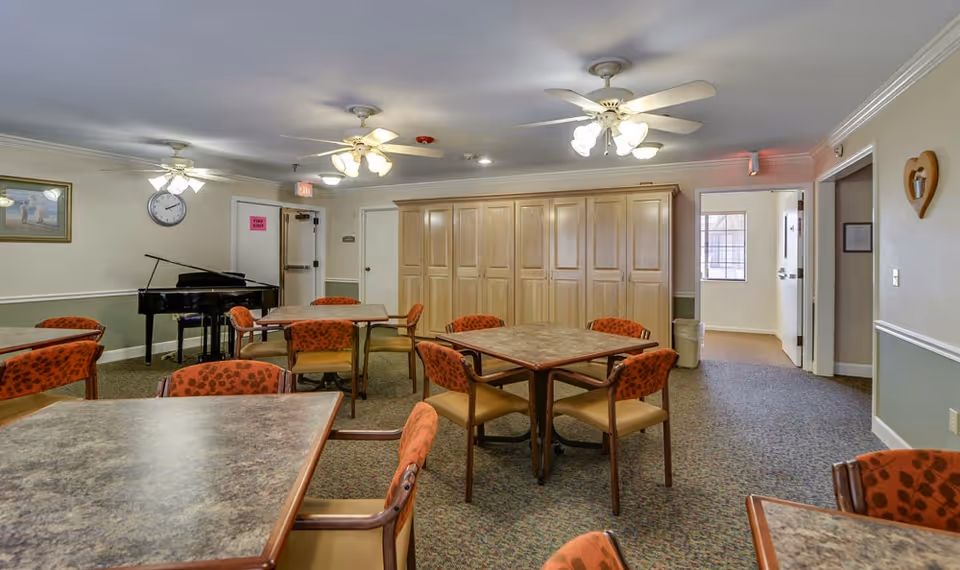 Community dining/activity room with multiple square tables and orange-patterned chairs, a black piano, ceiling fans, and large wooden storage cabinets.