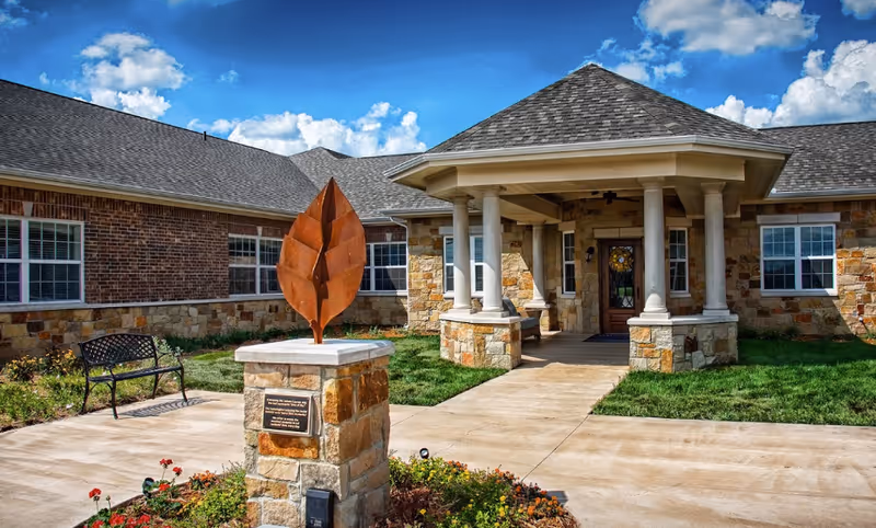 Front entrance of a memory care building with stone columns, a paved walkway, a metal leaf sculpture on a pedestal, a bench, and landscaped grounds under a blue sky.