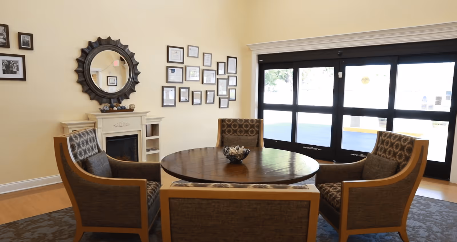 A cozy seating area with four patterned armchairs arranged around a round wooden table with a decorative bowl in the center. Behind the seating area is a white fireplace with a round decorative mirror above it and a wall adorned with framed certificates and photos. Large windows with black frames allow natural light to fill the room.
