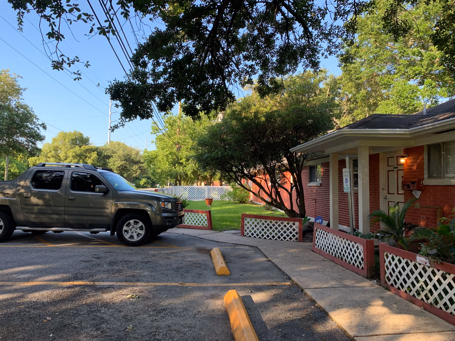 Parking area outside a single-story brick building with white lattice fencing and a small garden. A gray pickup truck is parked in the lot, and trees provide shade over the area.