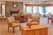 A cozy living room area with several cushioned armchairs arranged around wooden tables. There is a stone fireplace with a clock above it, a wooden cabinet with a vase of sunflowers, large windows with valances letting in natural light, and a potted plant near the windows.