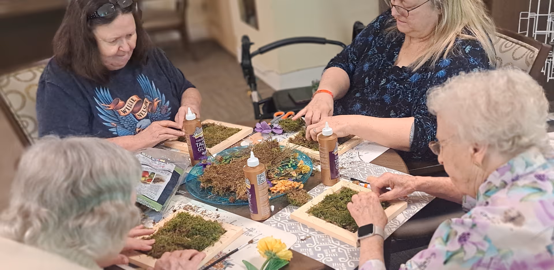 Four elderly women sitting around a table engaged in a craft activity, arranging moss and other natural materials on wooden frames with bottles of tacky glue on the table.