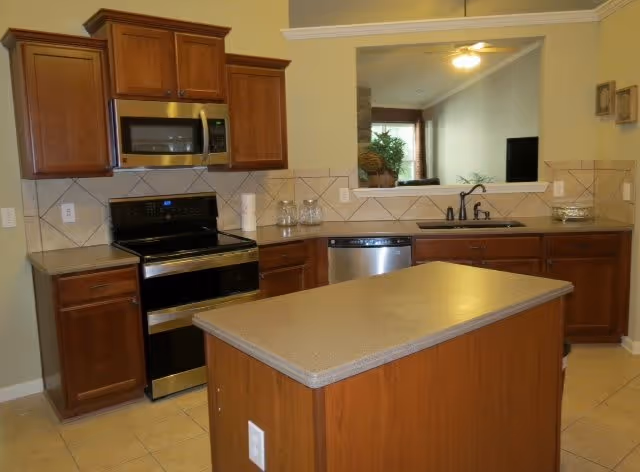 A kitchen with wooden cabinets, a stainless steel microwave above a black and stainless steel stove, a dishwasher, a sink with a black faucet, and a central island with a beige countertop. The kitchen has tiled flooring and a pass-through window looking into a living room area with a ceiling fan and a television.