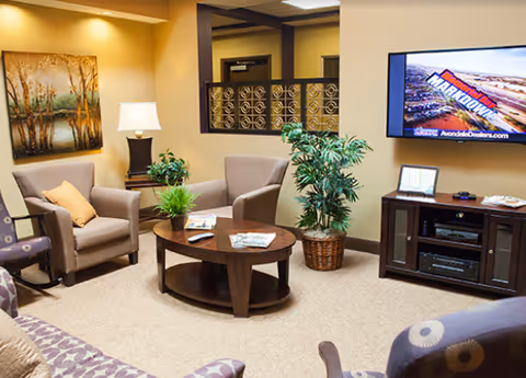 A cozy living room area with beige walls and carpet. The room features a wooden coffee table with magazines and a remote control, surrounded by several upholstered chairs and a sofa. There is a potted plant next to the coffee table and another larger plant in a wicker basket near the wall. A table lamp sits on a side table next to one of the chairs. A flat-screen TV is mounted on the wall above a dark wooden media console. A framed painting of trees hangs on the wall, and a decorative metal railing is visible in the background.
