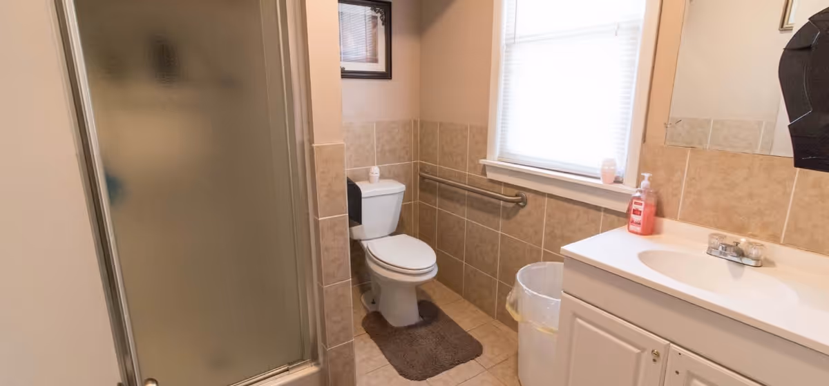 A clean bathroom with beige tiled walls and floor, featuring a white toilet with a dark brown rug in front, a frosted glass shower door on the left, a white sink with a countertop and a soap dispenser on the right, a trash can next to the sink, a window with a white blind above the toilet, and a metal grab bar on the wall beside the toilet.