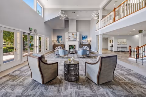 Spacious and bright senior living facility common area with high ceilings, large windows, and modern furnishings. The room features a seating arrangement with armchairs and a small round table in the foreground, additional chairs and sofas near a fireplace with artwork on the walls, and a second-floor balcony overlooking the space. Natural light fills the room through glass doors leading outside.