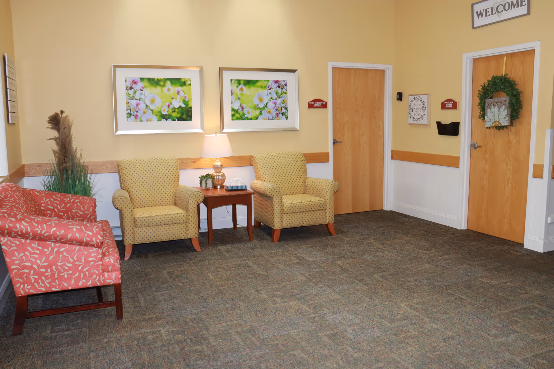 A waiting area with two yellow patterned armchairs and two red patterned chairs arranged around a small wooden table with a lamp and decorative plants. The walls are painted yellow with white wainscoting and decorated with two framed floral pictures. There are two wooden doors, one with a green wreath and a welcome sign above it. A 'WELCOME' sign is mounted on the wall above the door with the wreath.
