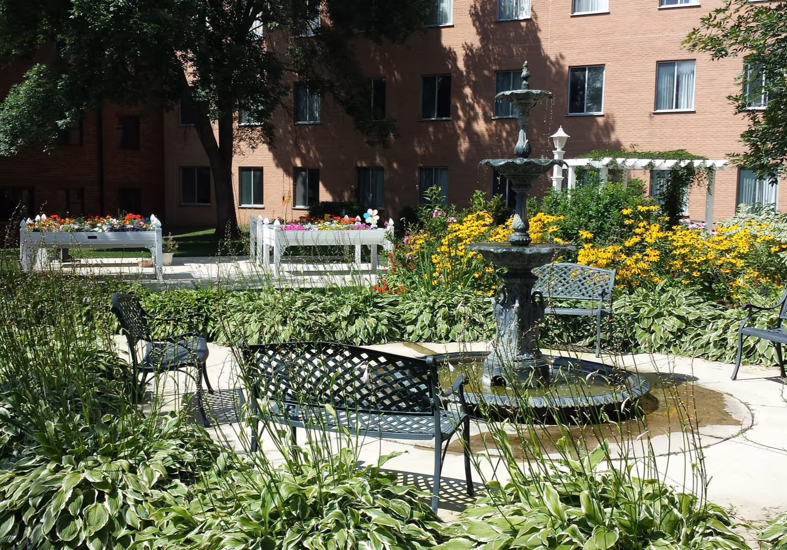 Outdoor garden area with a multi-tiered water fountain surrounded by metal benches and lush greenery. There are colorful flowers and raised flower beds in the background, with a brick building and windows visible behind the garden.