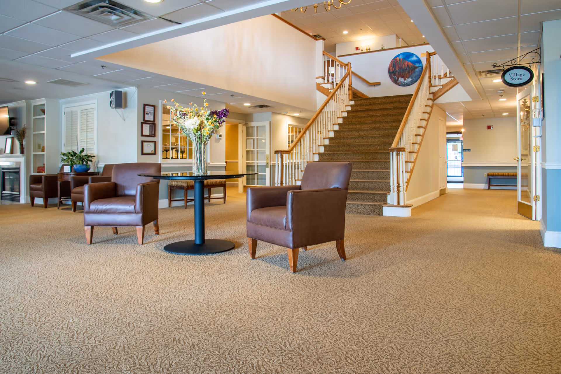 A spacious interior common area with beige carpet, two brown leather armchairs, and a round table with a vase of flowers. In the background, there is a staircase with wooden handrails leading to an upper floor, a fireplace with a TV above it, and a sign hanging from the ceiling that reads 'Village Store'.