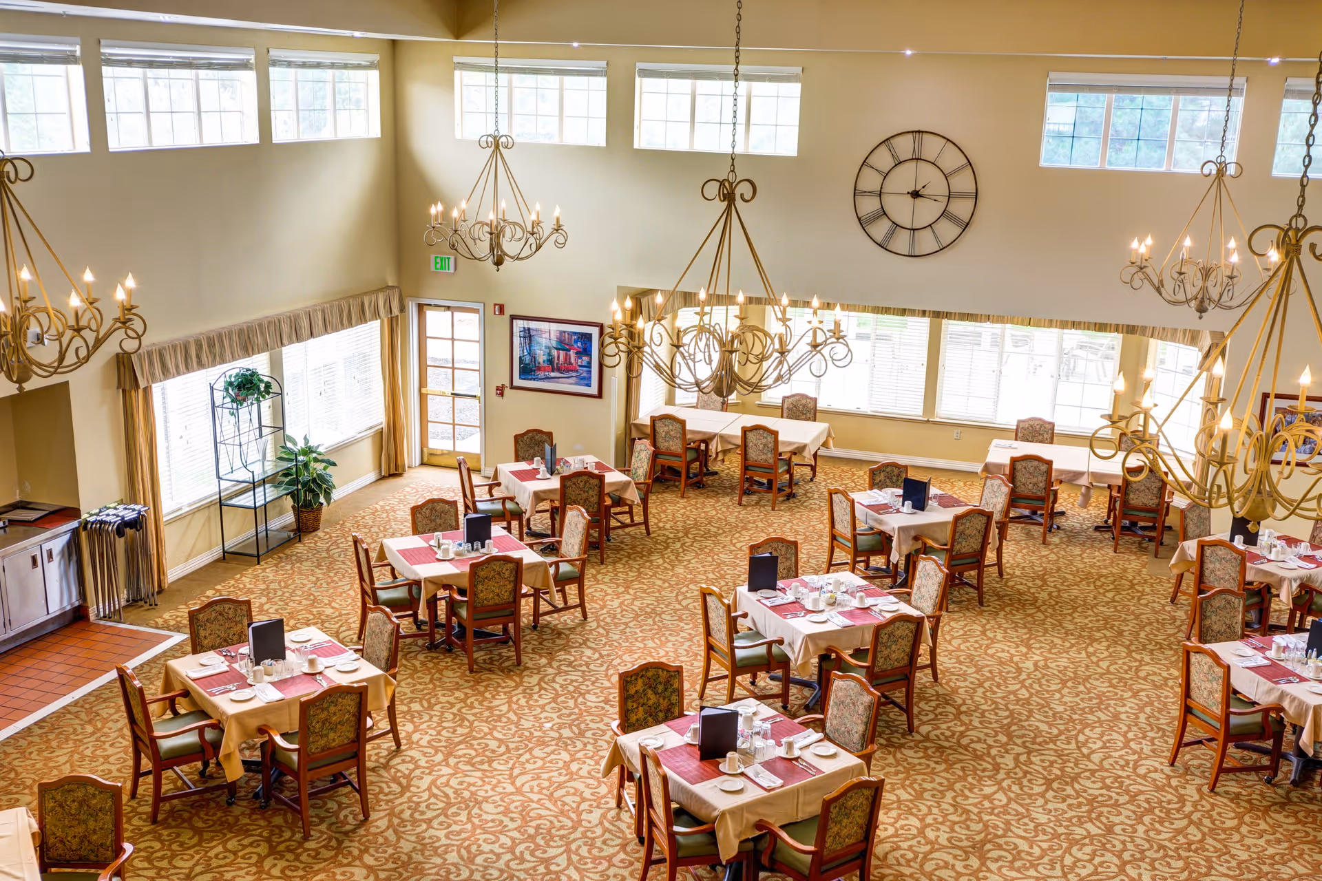 A spacious dining room with multiple tables covered in beige tablecloths and red placemats, each set with plates, cups, and menus. The room has large windows letting in natural light, ornate chandeliers hanging from the ceiling, a large wall clock, and patterned carpet flooring.