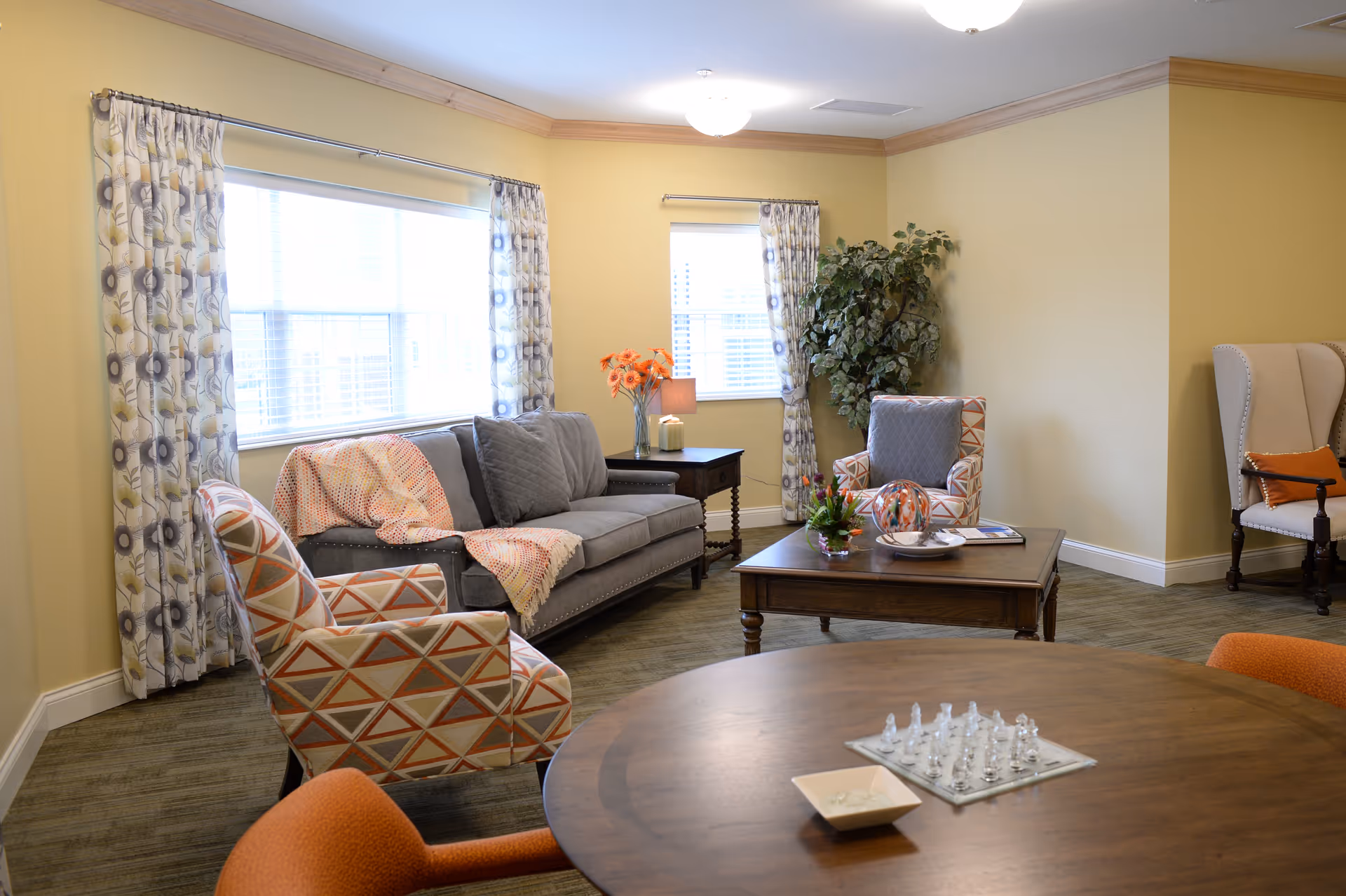 A cozy living room with a gray sofa adorned with a colorful throw blanket and pillows, two patterned armchairs, a wooden coffee table with decorative items, a side table with a lamp and flowers, and a round wooden table with a glass chess set. The room has yellow walls, large windows with floral curtains, and a potted plant in the corner.