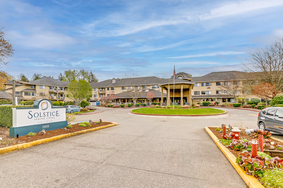 Front exterior of Solstice Senior Living at Bellingham showing the entrance canopy, circular driveway, landscaping, and a facility sign in the foreground.