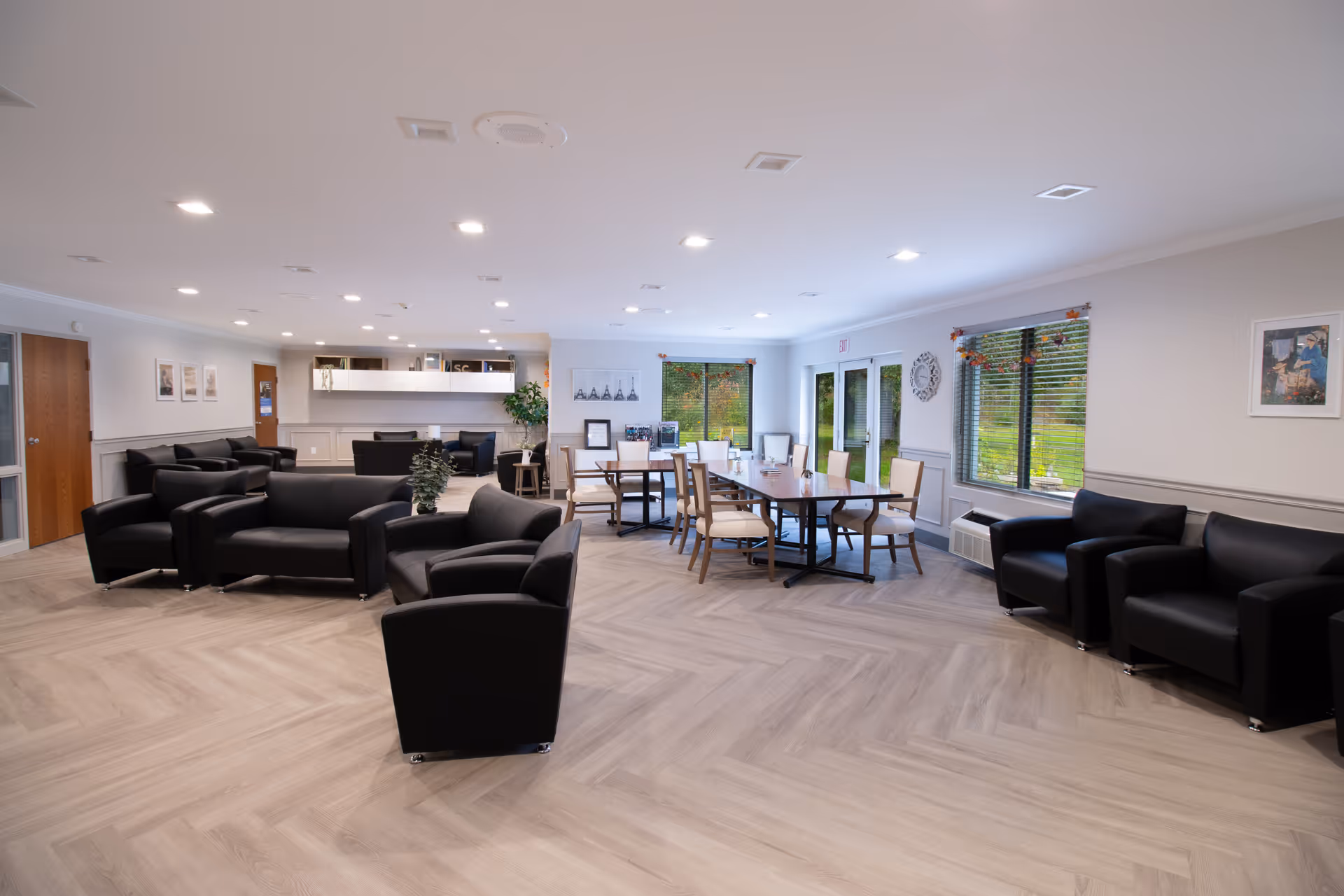 A spacious, well-lit common area in a senior living facility featuring multiple black leather armchairs and sofas arranged in seating groups, a long wooden table with chairs near large windows, light wood flooring, and neutral-colored walls with framed artwork and decorations.