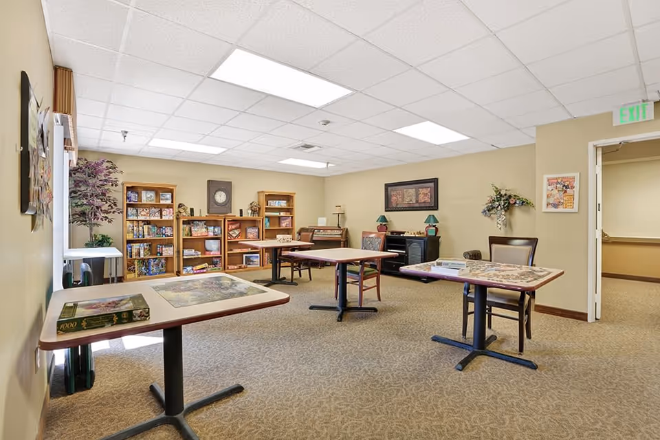 A well-lit activity room with beige walls and carpeted floor featuring several tables with puzzles on them, wooden bookshelves filled with board games and books, a clock on the wall, a piano, and chairs arranged around the tables. There is a plant in the corner and framed artwork on the walls.