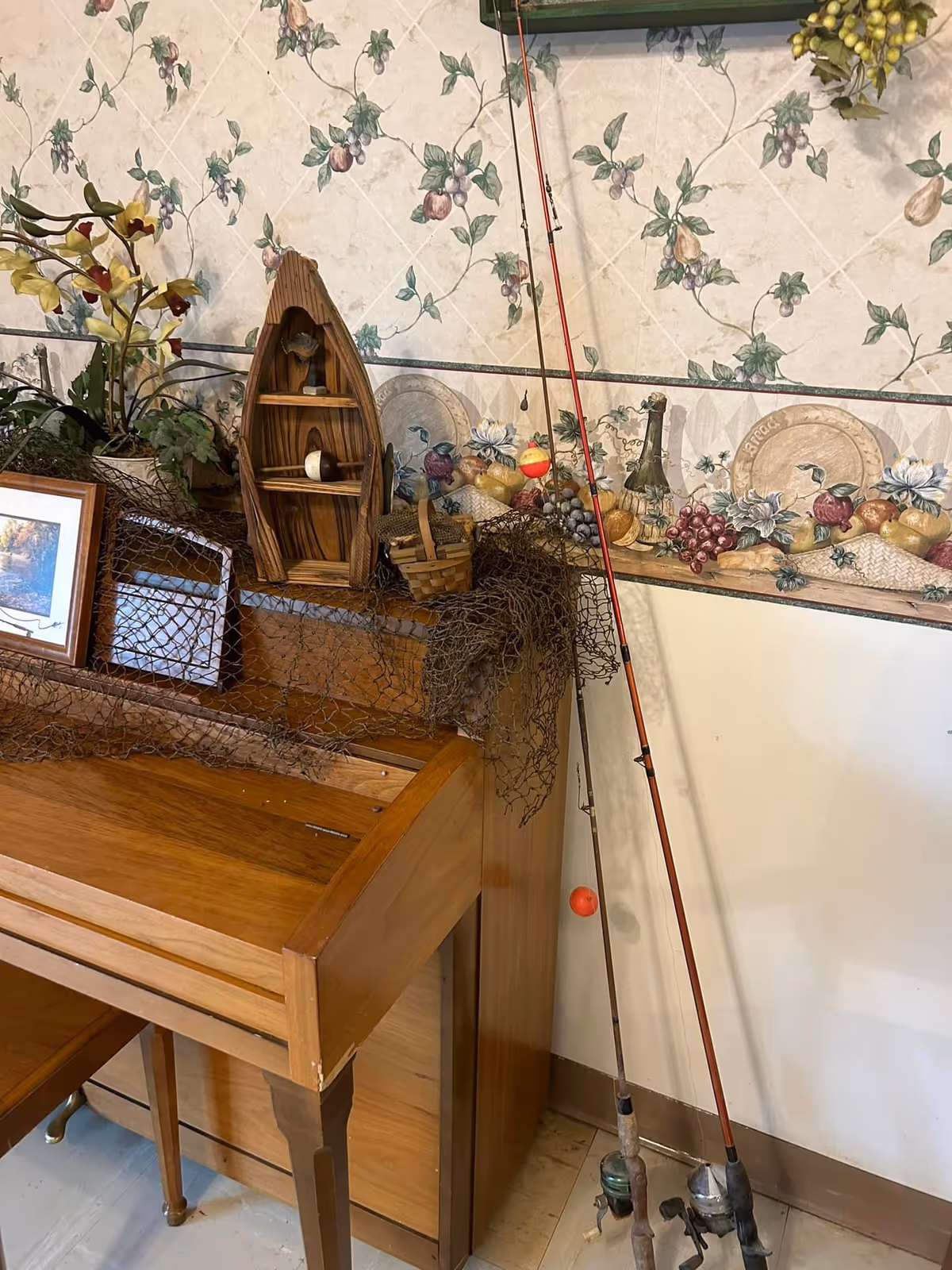 A corner of a room with a wooden roll-top desk. On top of the desk are decorative items including a small wooden boat-shaped shelf, a fishing net, a small basket, and a framed picture. Two fishing rods lean against the wall next to the desk. The wall has floral and fruit patterned wallpaper with a border featuring plates and fruit.