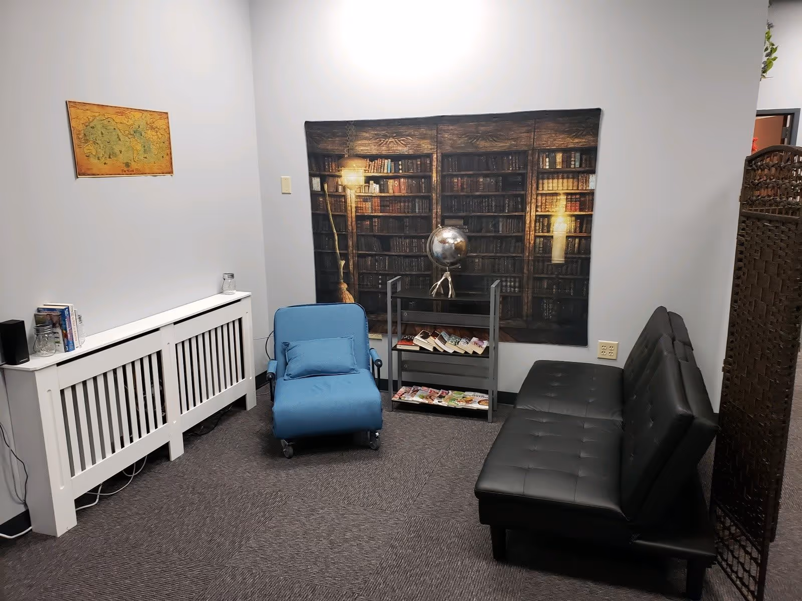 A small seating area in a room with a black leather couch, a blue cushioned chair, a small bookshelf with books and magazines, and a decorative wall hanging depicting a bookshelf with old books and a globe. There is also a map on the adjacent wall and a white radiator cover with some items on top.