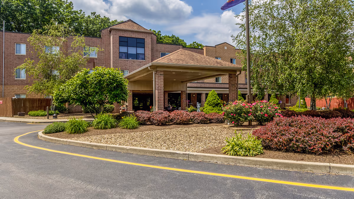 Front exterior view of a three-story brick senior living community building with a covered entrance, surrounded by landscaped bushes, trees, and flowers under a partly cloudy sky.