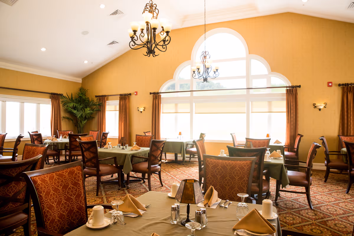 Bright, upscale dining room with multiple set tables, chandeliers, large arched windows, and upholstered chairs.