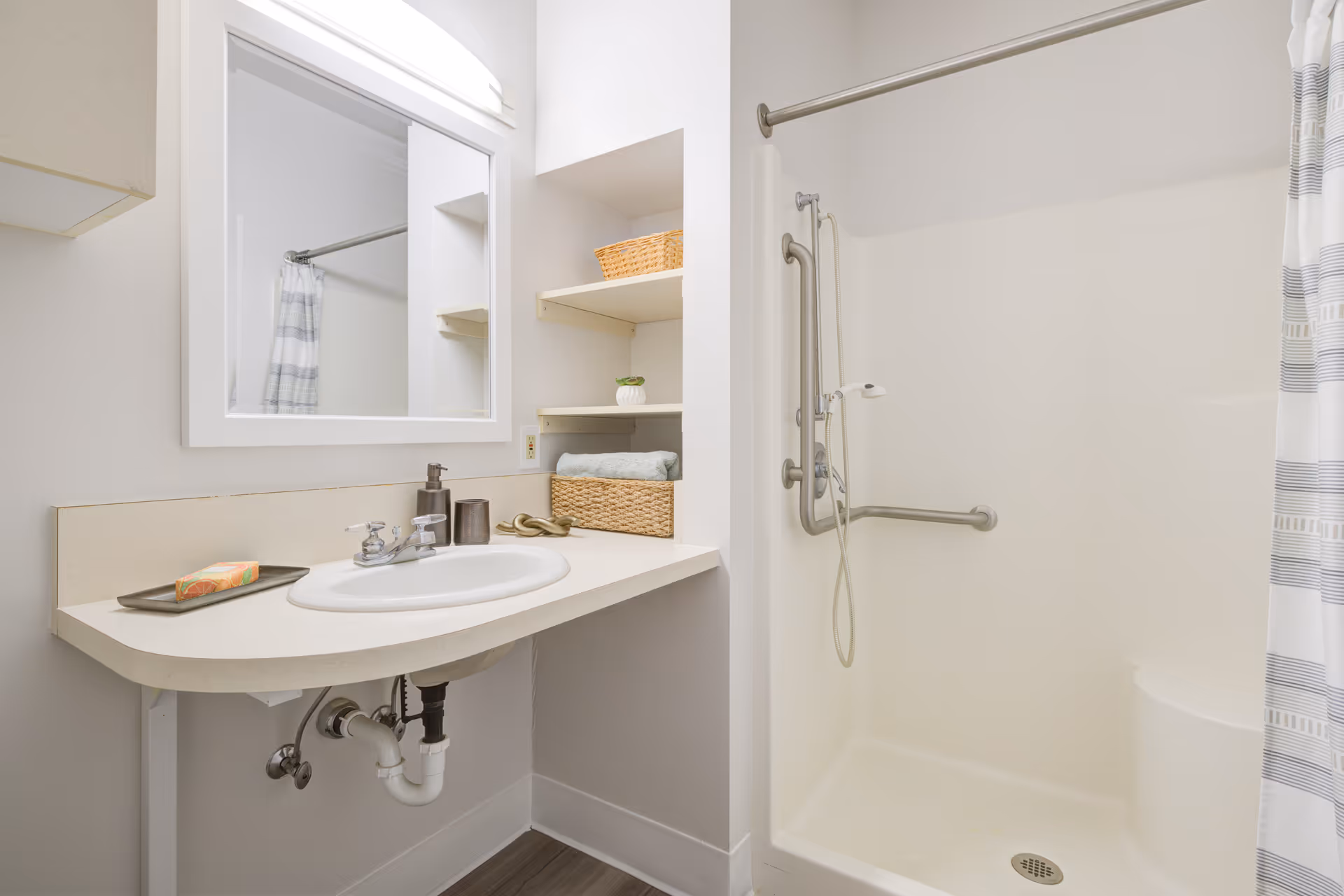 A bright accessible bathroom with a sink and mirror, open shelving with baskets, and a walk-in shower with grab bars and a striped curtain.