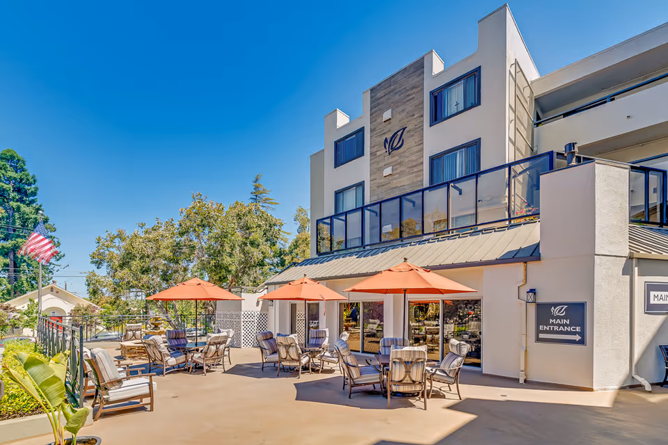 Outdoor patio with tables, umbrellas, and seating in front of a multi-story senior living building marked 'Main Entrance'.