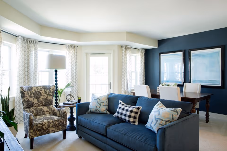 A bright and cozy living room area with a blue sofa adorned with patterned pillows, a floral armchair, a round side table with a lamp and decorative items, and a dining table with white chairs against a dark blue accent wall featuring two framed abstract paintings.