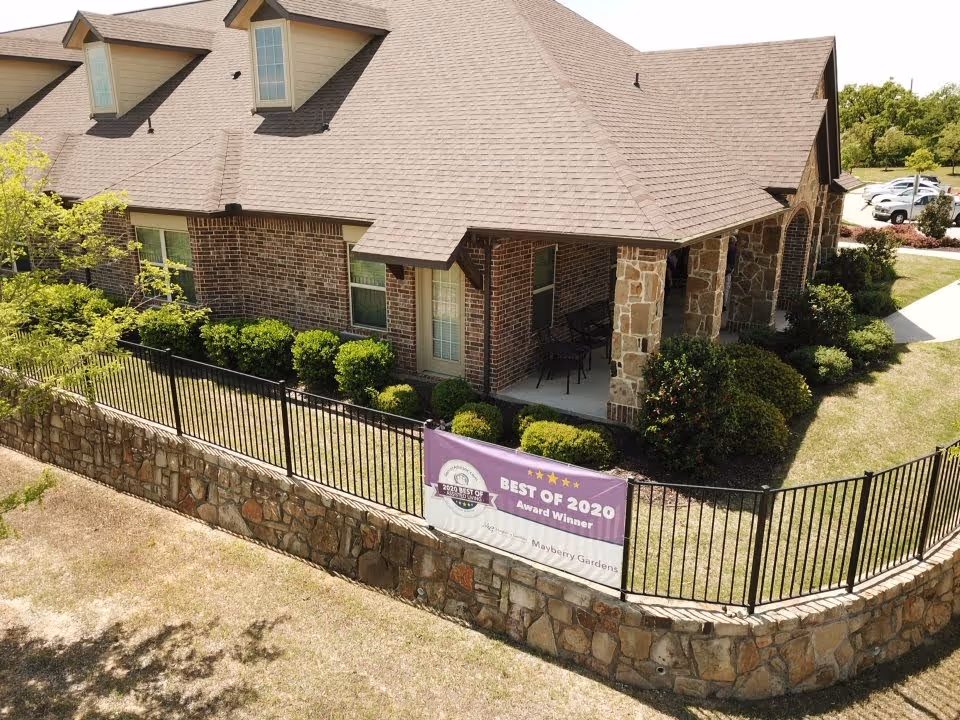 Front exterior of a brick and stone assisted living building with a fenced yard and a 'Best of 2020' banner.