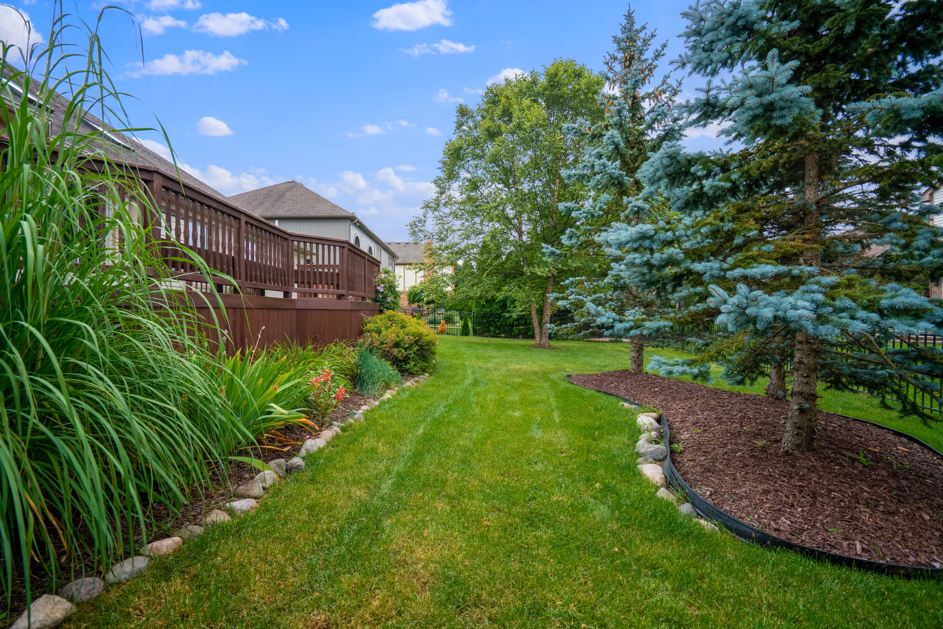 A well-maintained outdoor garden area with green grass, various plants, and trees. There is a wooden deck on the left side and a mulched area with evergreen trees on the right. The sky is blue with scattered clouds.