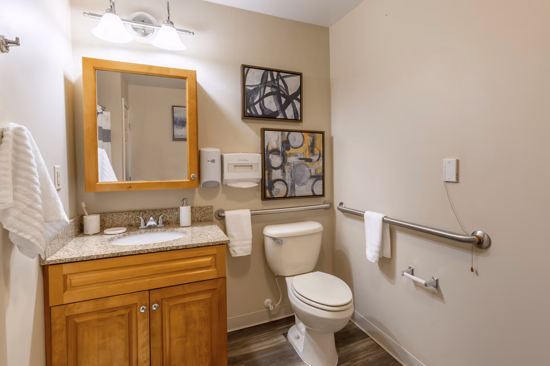 A clean and well-lit bathroom with a wooden vanity featuring a granite countertop and a sink. Above the vanity is a wooden framed mirror with two light fixtures. To the right of the vanity is a white toilet with safety grab bars on the wall beside it. Two abstract paintings hang on the wall above the toilet. White towels are hanging on the grab bars and a towel rack on the left wall. The floor has wood-like tiles.