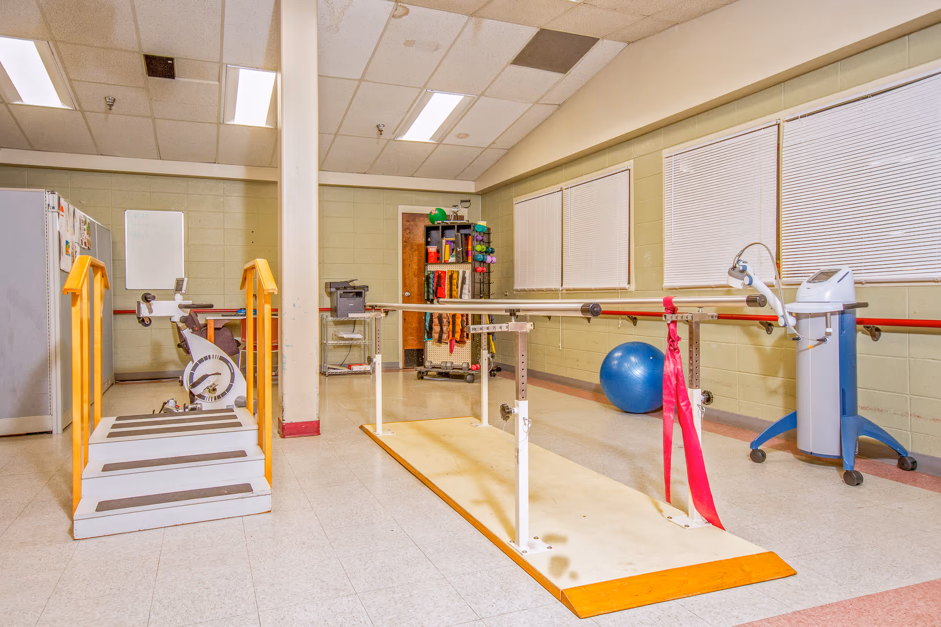 Therapy/rehab room containing parallel bars, a set of steps with handrails, an exercise bike, therapy ball, and other equipment.