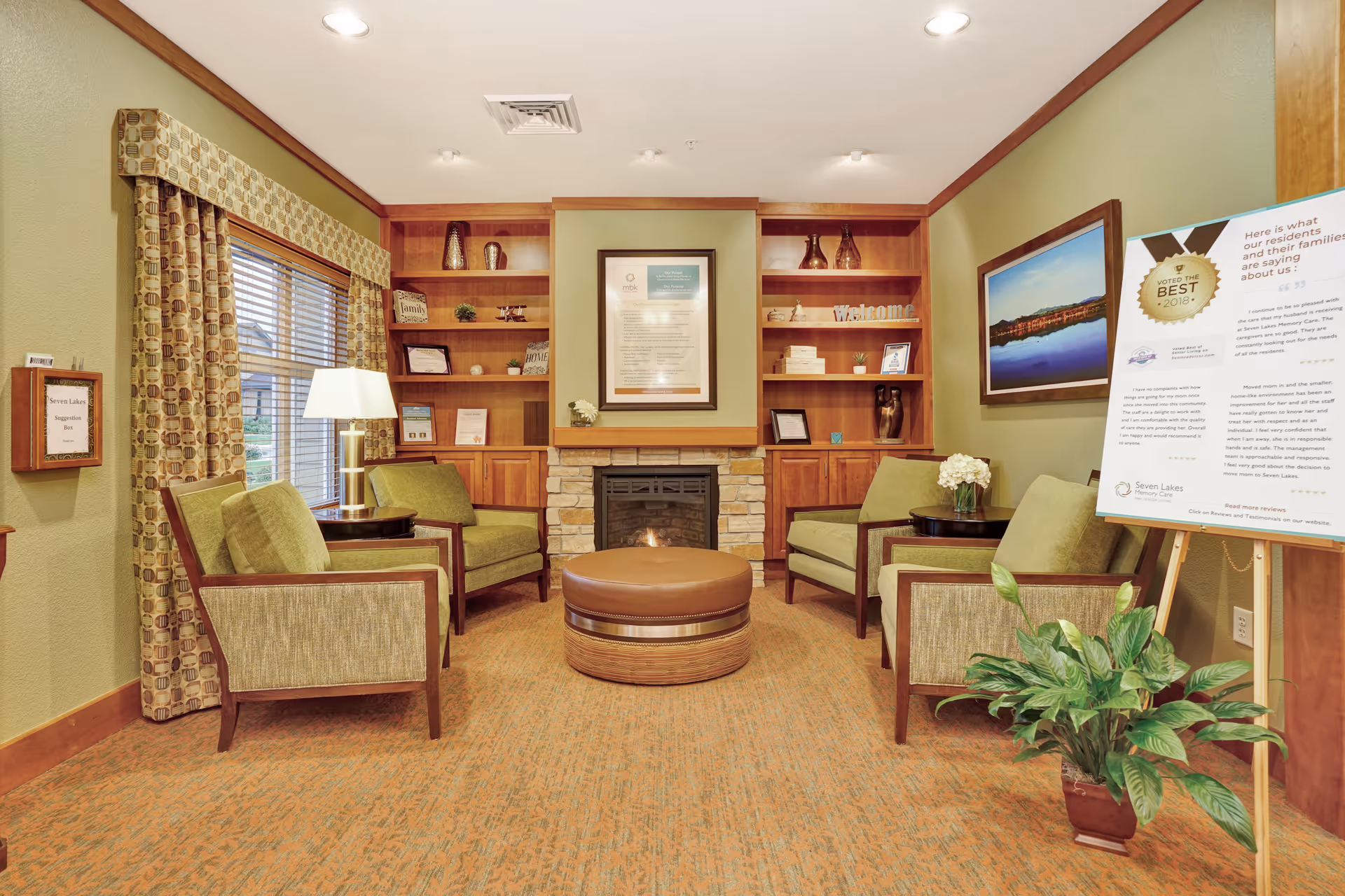 A cozy sitting area in a memory care facility with four green upholstered armchairs arranged around a round brown ottoman. The room features built-in wooden shelves with decorative items, a stone fireplace, a large window with patterned curtains, a floor lamp, a framed picture on the wall, and a large informational sign on an easel. There is also a potted plant on the floor.