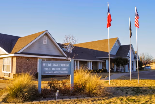 Exterior view of Wildflower House, a licensed assisted living facility located at 706 Red Coat Drive. The building is a single-story structure with a brick and siding facade, surrounded by grass and ornamental grasses. Three flagpoles with flags are positioned near the entrance.